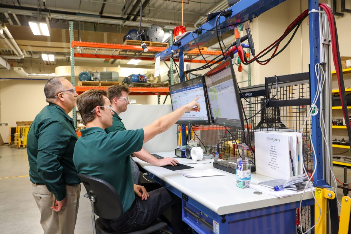 Three researchers looking at 2 screens. One of them is pointing at one screen to specific data. The are standing at a lab bench